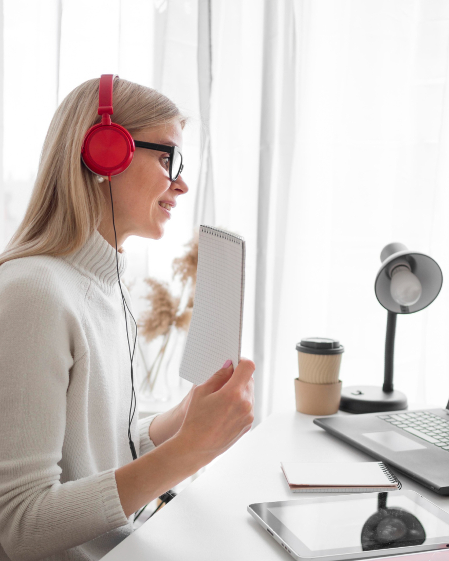 Afbeelding: Een vrouw met een rode koptelefoon en een bril maakt aantekeningen terwijl ze aan een bureau bij een laptop zit