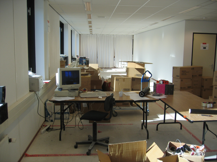 Afbeelding: Vroege kantoorruimte van iBOOD met dozen, eenvoudige bureaus en een oude computeropstelling, als beeld van de pioniersfase van het bedrijf